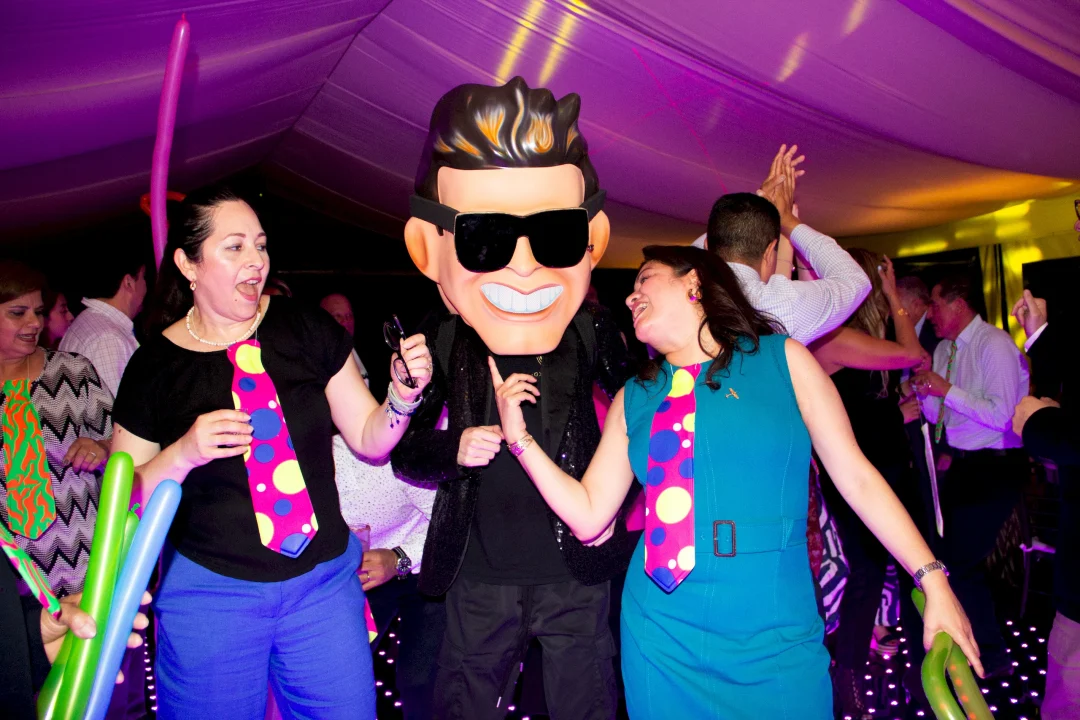 Invitados divirtiéndose en una pista de baile LED durante un evento animado por Axis Live. Dos mujeres bailan entusiasmadas junto a un animador que lleva una gran cabeza de personaje de caricatura con gafas de sol y traje de lentejuelas. Las mujeres visten corbatas de utilería de colores neón y la escena está llena de energía bajo una iluminación ambiental púrpura. Esta foto muestra la experiencia de animación temática y la diversión interactiva que ofrece Axis Live para bodas de lujo y eventos corporativos premium.