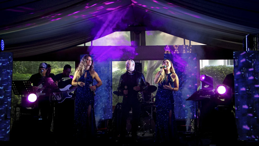Presentación elegante de Axis Live en un escenario íntimo bajo una carpa: dos vocalistas femeninas con vestidos de gala azul noche con lentejuelas y un cantante masculino con saco brillante, interpretando un repertorio de baladas y oldies. La escena cuenta con músicos en vivo (guitarrista y bajista), pantallas LED verticales con gráficos abstractos, iluminación ambiental en tonos violeta y el logotipo de "AXIS" iluminado al fondo, creando una atmósfera sofisticada para cenas exclusivas, bodas de lujo, xv años espectaculares y eventos corporativos.